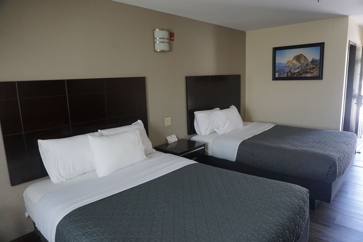 A hotel room with two beds, each with a dark headboard and white bedding. There is a nightstand with a phone and a wall-mounted light fixture. A framed picture of a mountain landscape is on the wall.