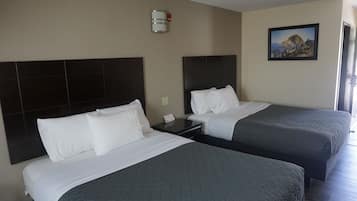 A hotel room with two beds, each with a dark headboard and white bedding. There is a nightstand with a phone and a wall-mounted light fixture. A framed picture of a mountain landscape is on the wall.