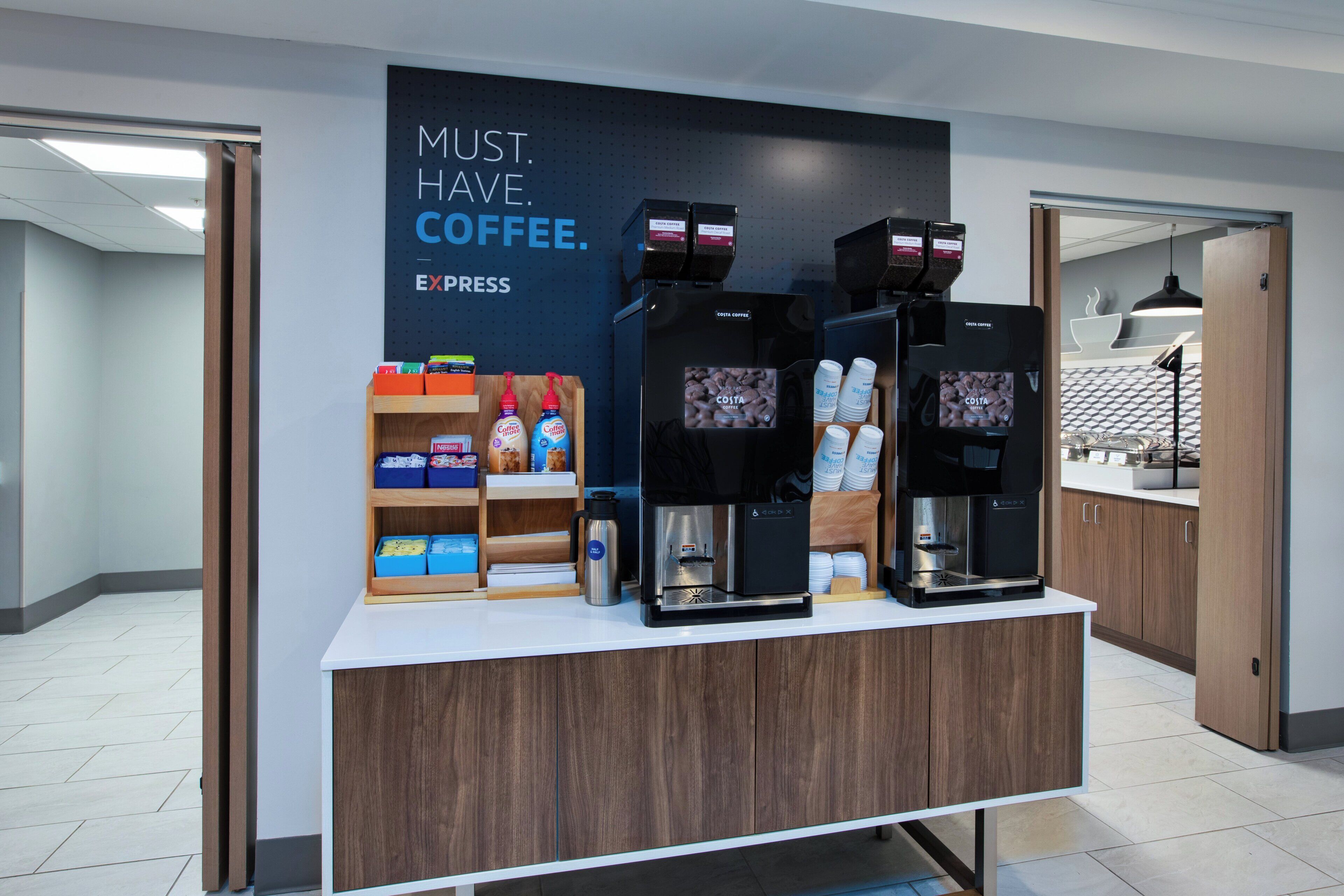 A coffee station with three espresso machines, a display of coffee pods, and a sign that reads "MUST. HAVE. COFFEE."