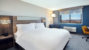 A hotel room with a large bed, a nightstand, a desk, a chair, a window with a city view, and a wall-mounted lamp. - Holiday Inn Express Sault Ste. Marie by IHG (Sault Ste. Marie)