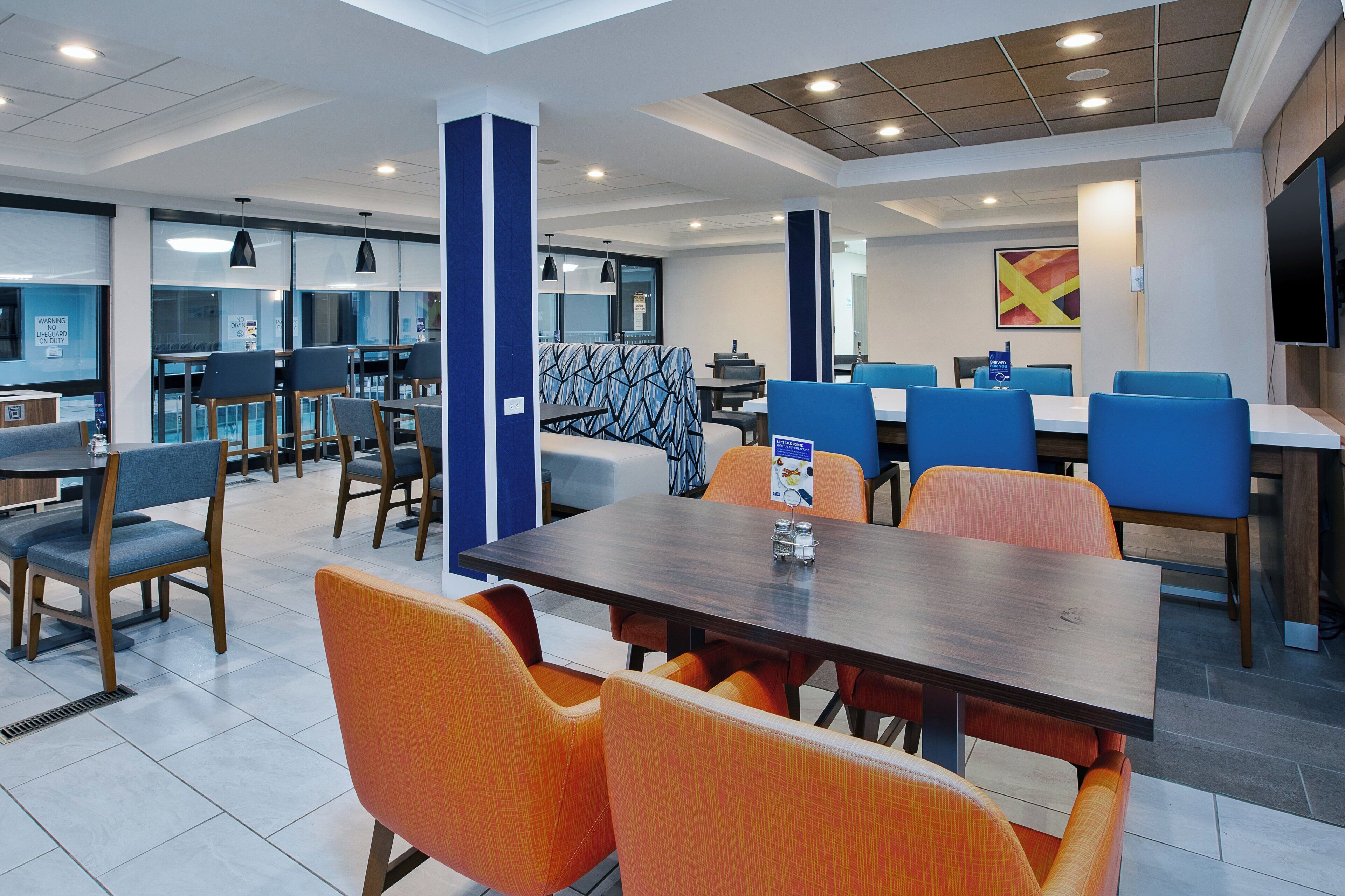 Un moderno vestíbulo de hotel con varias zonas de estar, incluyendo una mesa central con sillas naranjas y un sofá estampado, rodeado de sillas azules y naranjas.