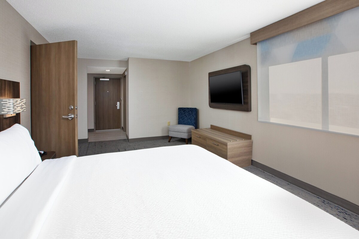 Ein Hotelzimmer mit einem großen Bett, einer Holzkommode, einem flachbildfernseher an der Wand und einer kleinen Sitzecke mit Sessel und Hocker.
