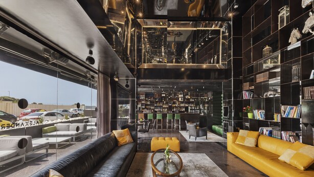 Un intérieur moderne avec un canapé en cuir noir, un pouf jaune et une table basse en verre. On y trouve également un coin bar avec une variété de bouteilles et une bibliothèque remplie de livres.