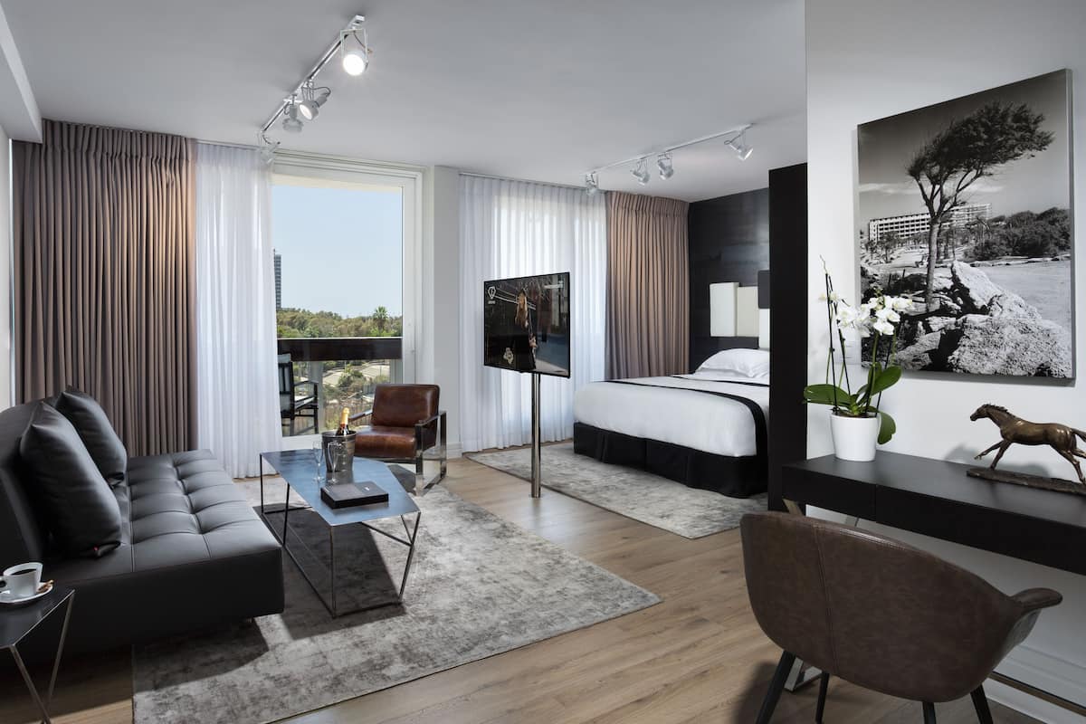 Een moderne hotelkamer met een bank, bureau, bed en televisie. Grote ramen bieden uitzicht op de stad.