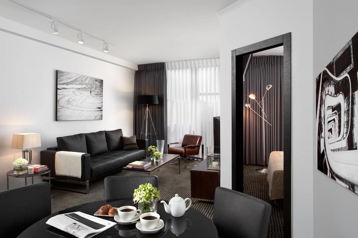 Een moderne hotelkamer met een zwarte leren bank, een ronde salontafel, een eettafel met kopjes en een theepot, een bruine lederen fauteuil en een groot abstract schilderij aan de muur.