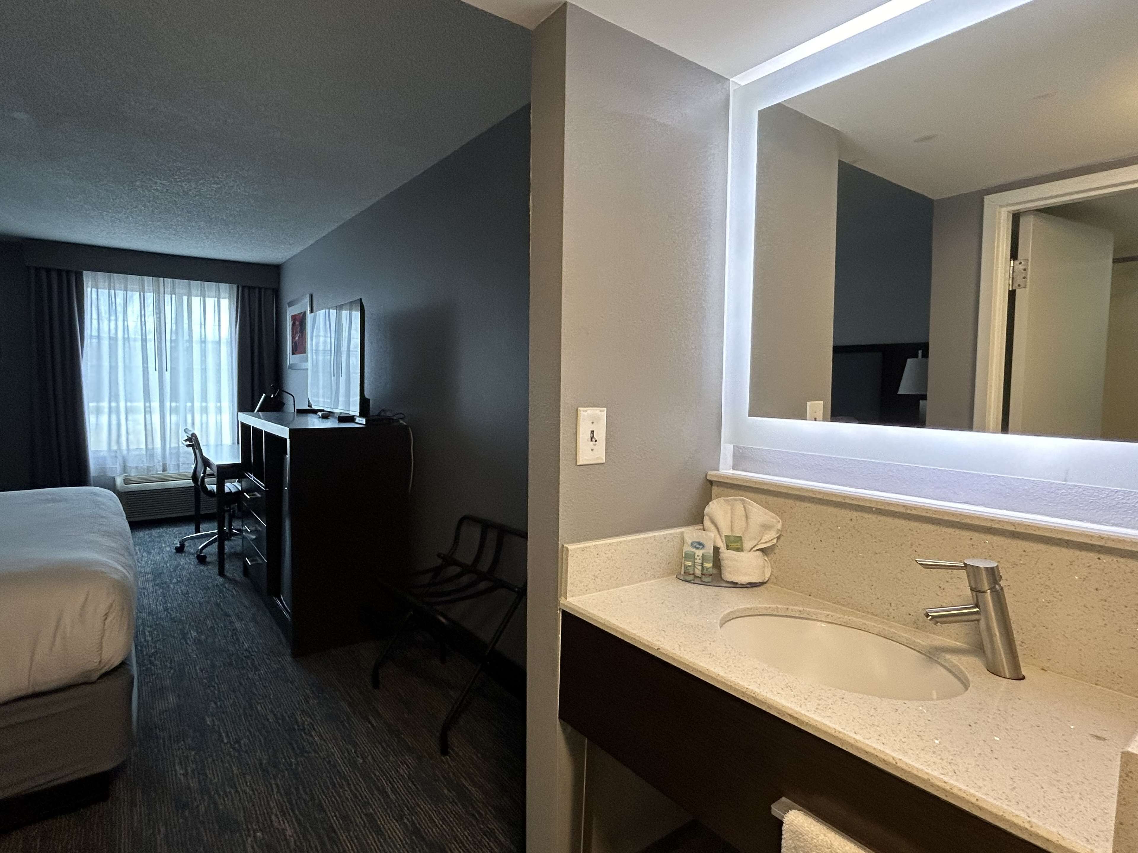A hotel room with a bed, a desk with a computer, a chair, a desk lamp, a window with curtains, and a bathroom with a sink and mirror.