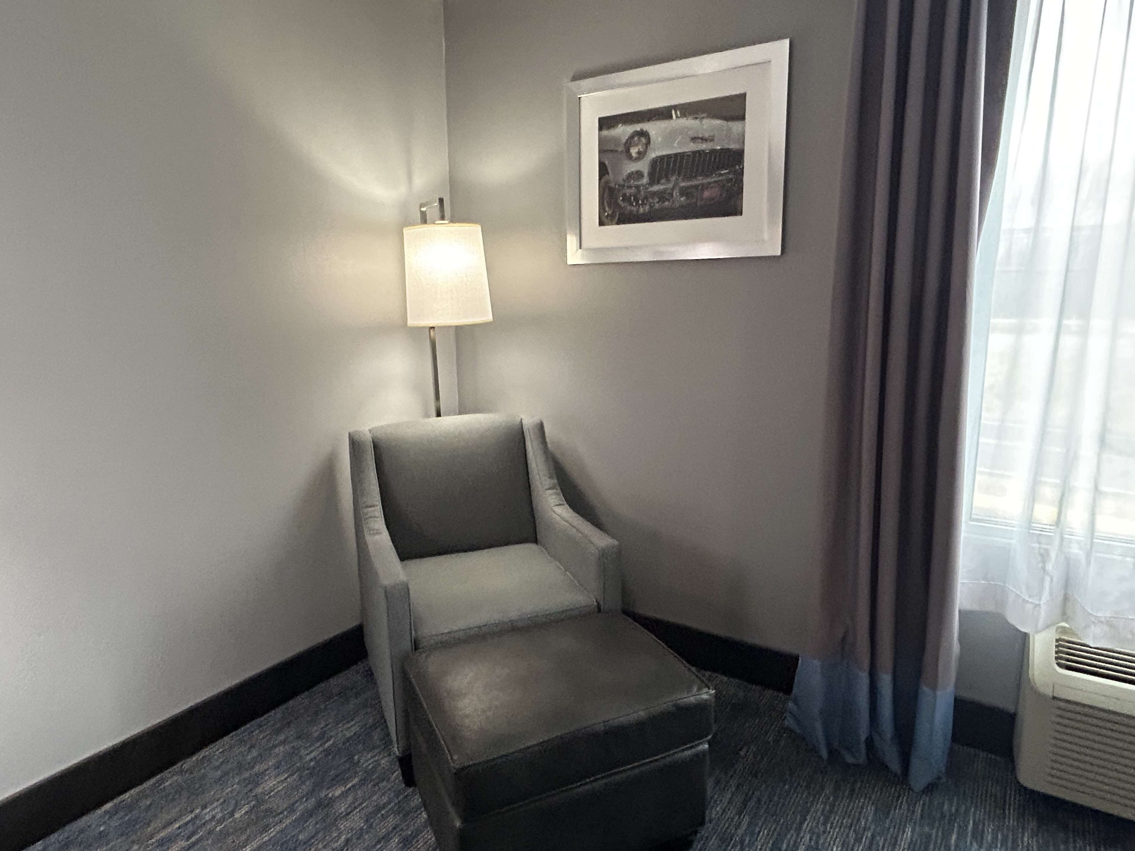 A hotel room with a grey armchair, a small ottoman, a framed picture on the wall, a lamp, and a window with curtains.
