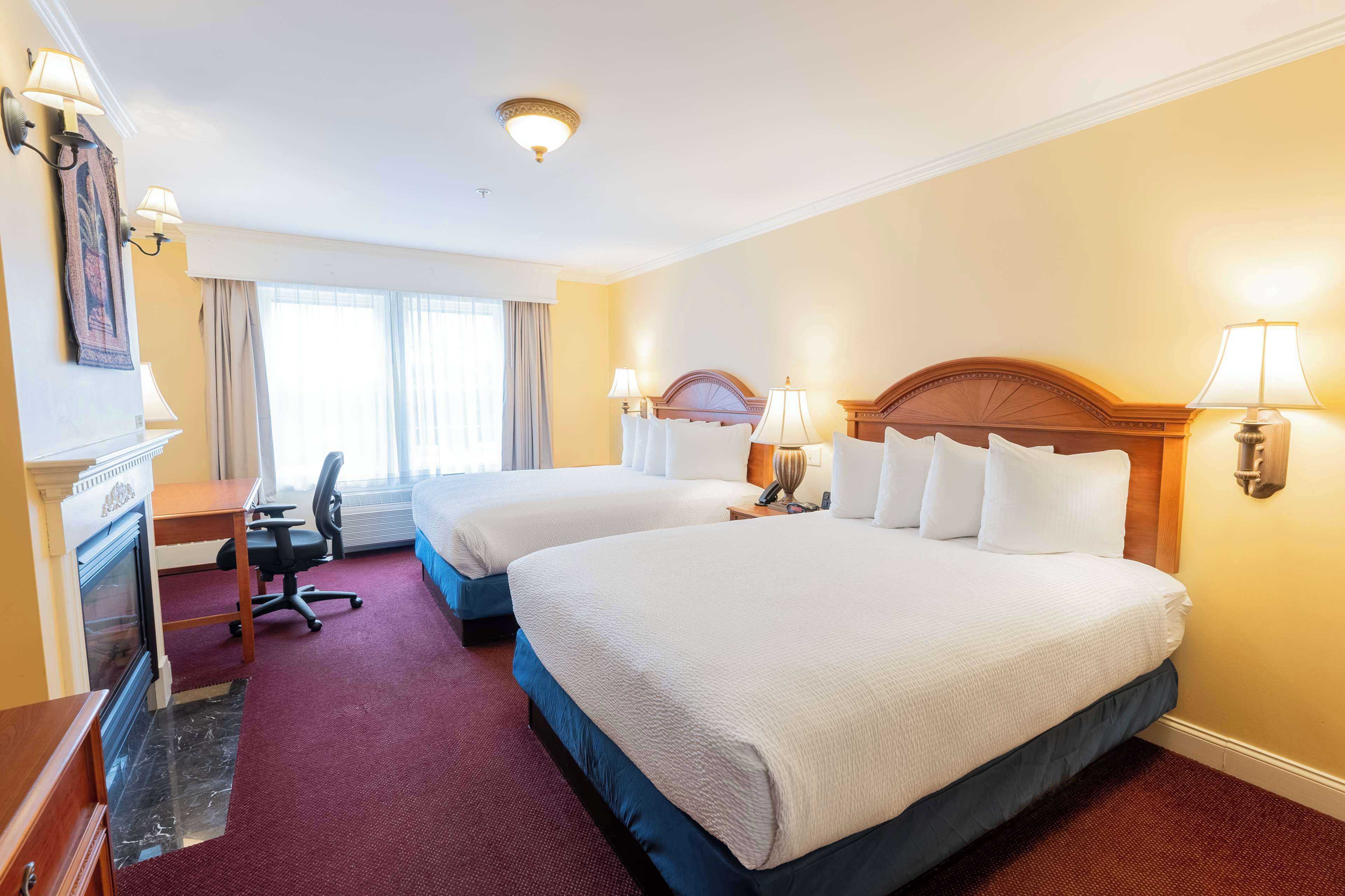 Une chambre d’hôtel avec deux lits, un bureau, une chaise, une cheminée et une grande fenêtre.