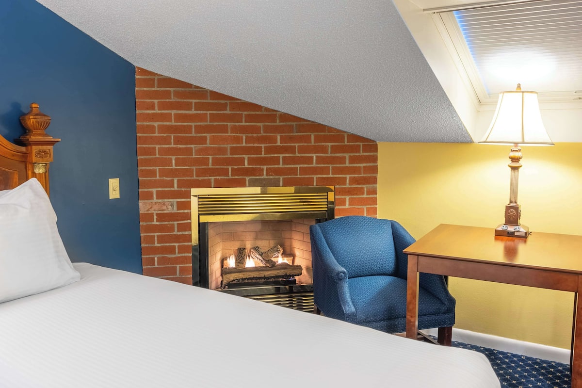 A hotel room with a brick fireplace, a bed with white linens, a blue armchair, and a wooden table with a lamp.