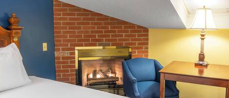 A hotel room with a brick fireplace, a bed with white linens, a blue armchair, and a wooden table with a lamp.