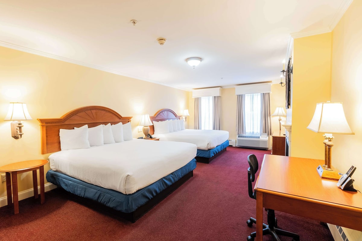 A hotel room with two beds, a desk, a chair, and a lamp.