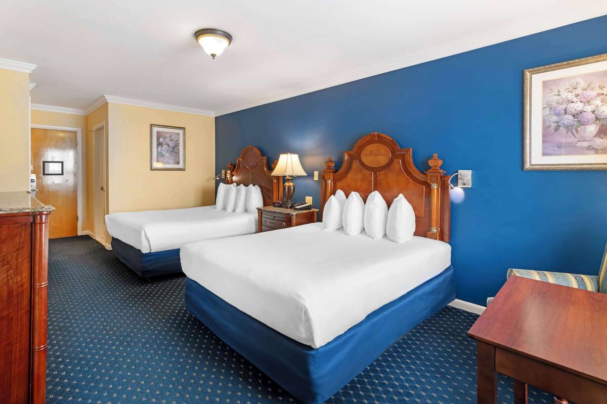 A hotel room with two beds, a wooden headboard, a nightstand with a lamp, a framed picture on the wall, and a blue carpet.