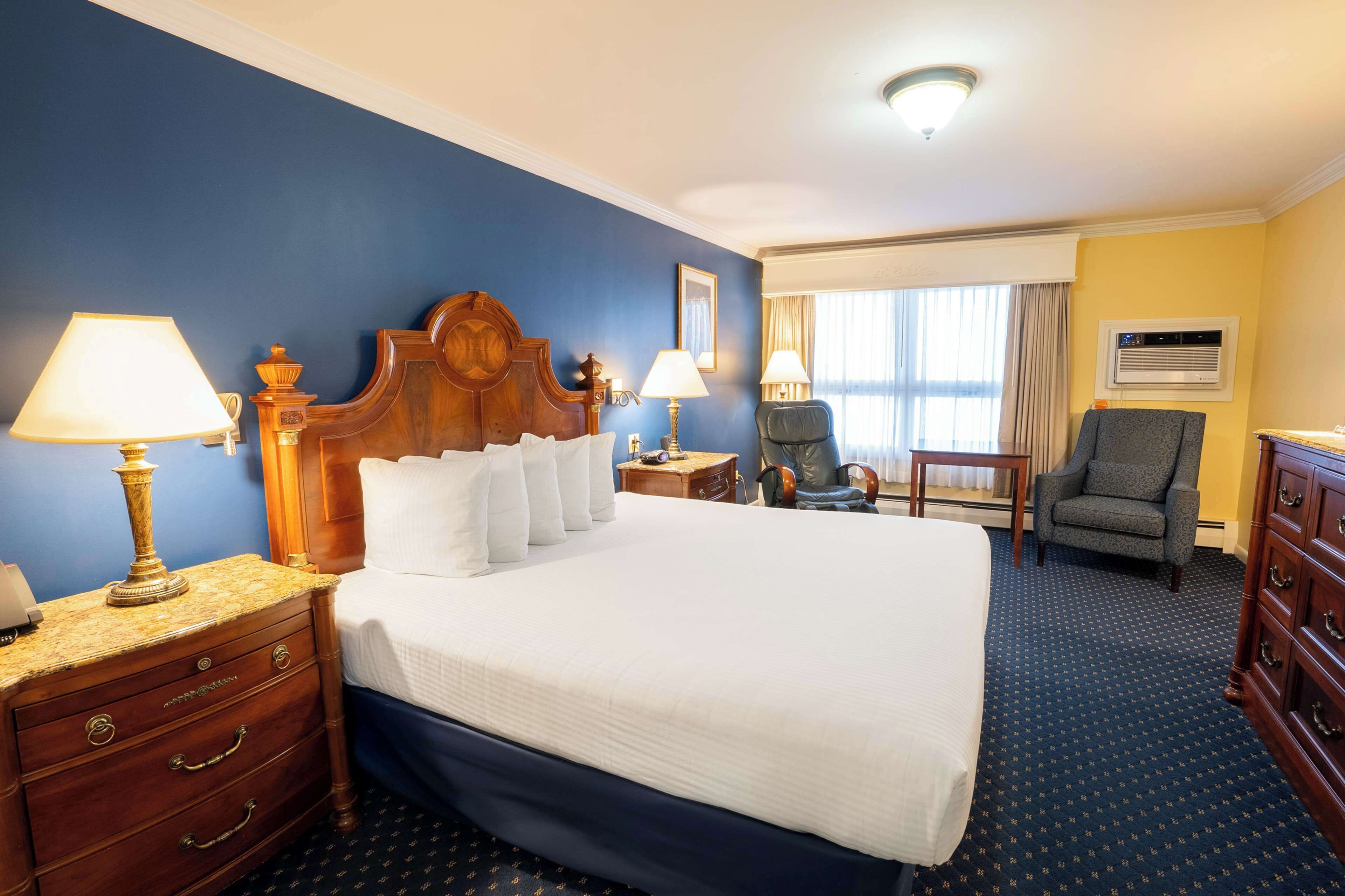 A hotel room with a large bed, two bedside tables with lamps, a chair, a dresser, and an air conditioning unit.