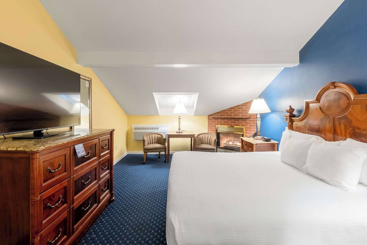 A hotel room with a bed, a TV on a dresser, a desk with a lamp, and a fireplace.
