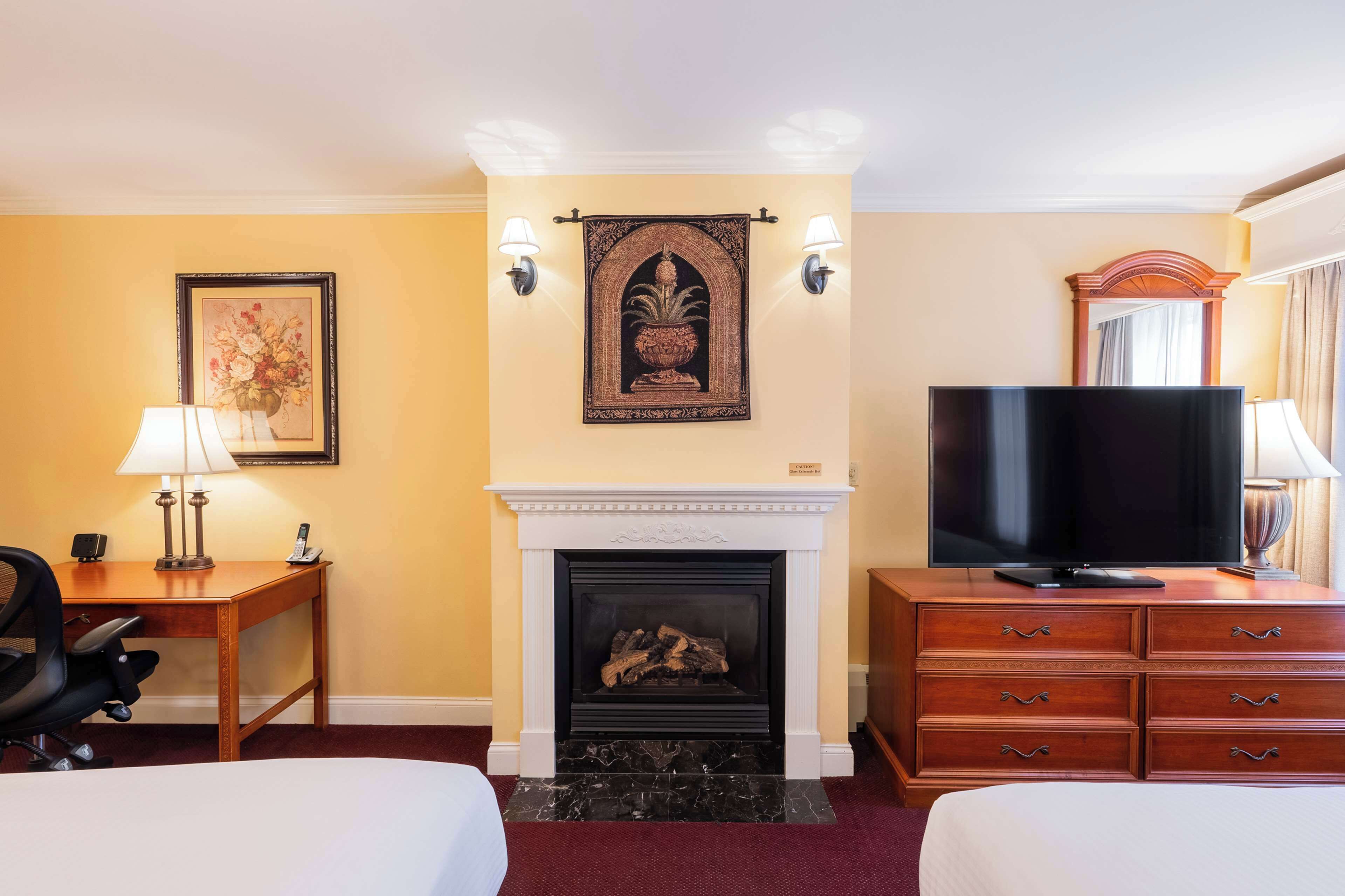 A hotel room with a fireplace, a television, a desk with a lamp, and a bed.