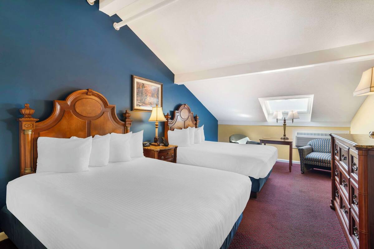 A hotel room with two large beds, a wooden headboard, a desk with a lamp, a chair, and a dresser.