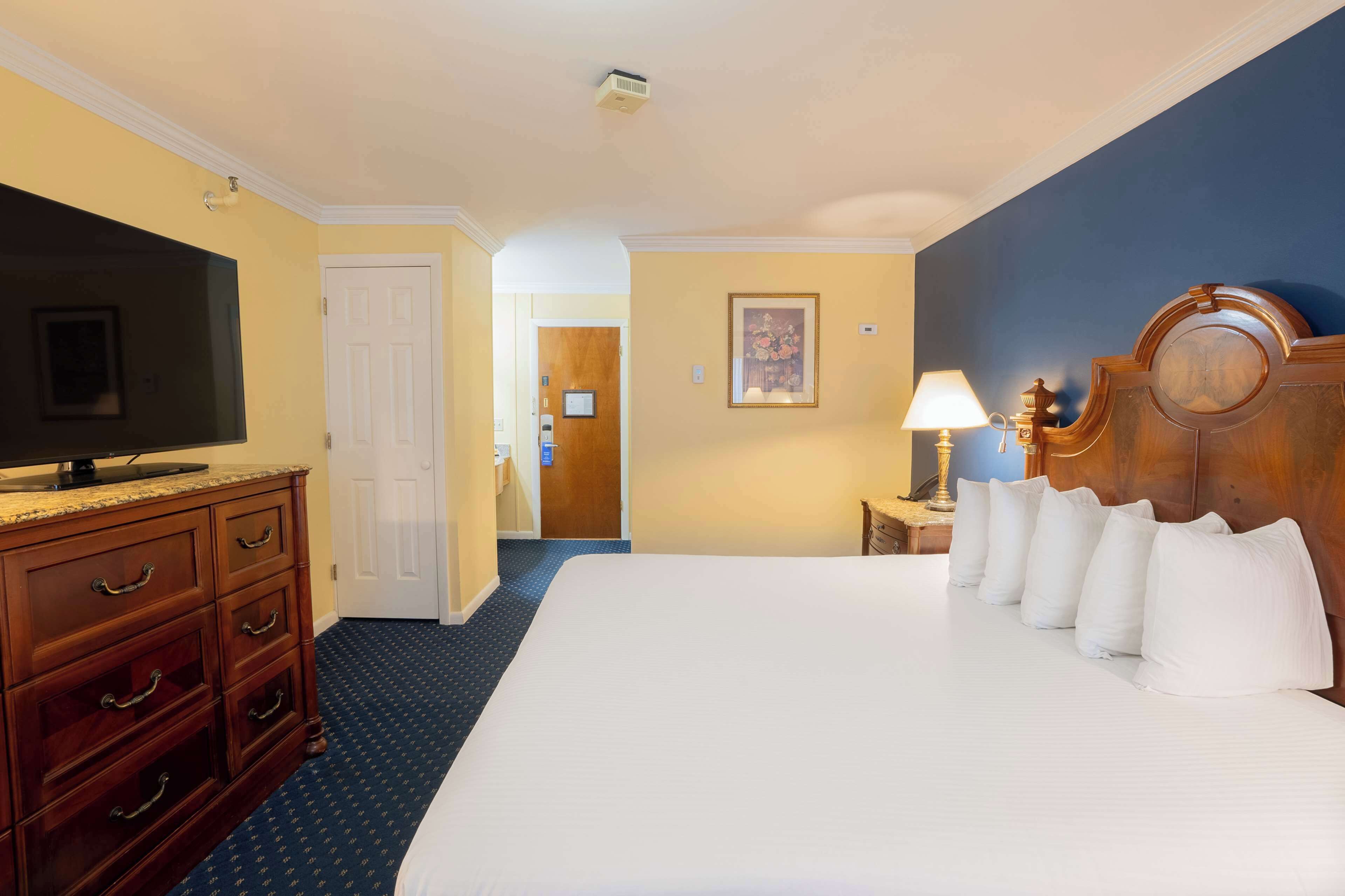 A hotel room with a large bed, a TV, a wooden dresser, and a nightstand with a lamp.