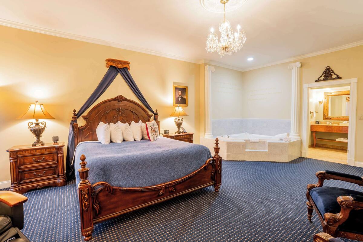 A bedroom with a four-poster bed, a sitting chair, a nightstand, a lamp, a large mirror, and a bathroom with a bathtub and sink.