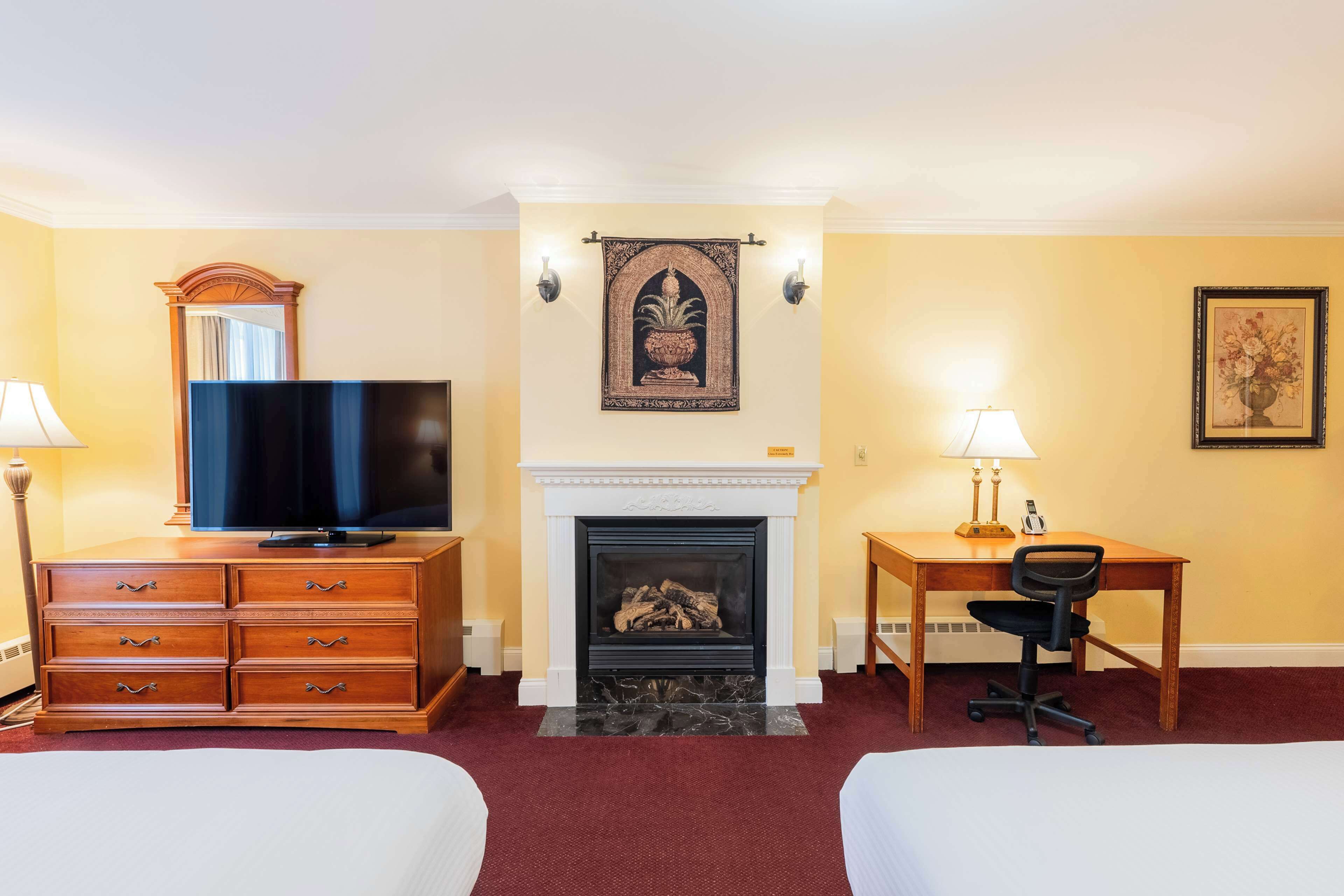 A hotel room with two beds, a television, a fireplace, a desk with a chair, and wall-mounted lamps.