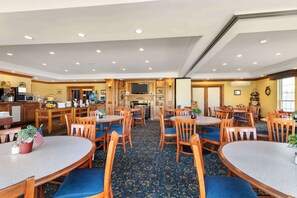 A hotel dining area with round tables and chairs, a kitchen area with a TV, and a fireplace.