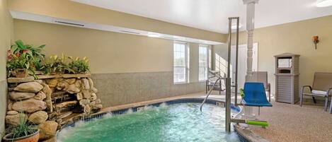 An indoor swimming pool with a stone wall and plants, a blue slide, and a seating area with chairs.