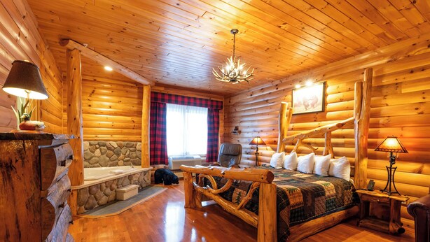A log cabin interior with a stone fireplace, a wooden bed, a stone bench, and a chandelier.