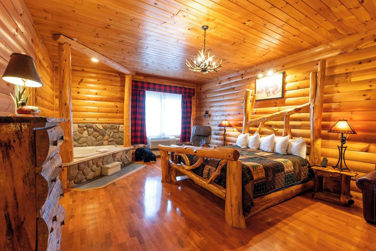 A log cabin interior with a stone fireplace, a wooden bed, a stone bench, and a chandelier.