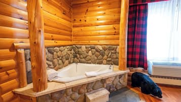 A wooden cabin interior with a stone wall, a wooden bathtub, and a black dog on the floor.