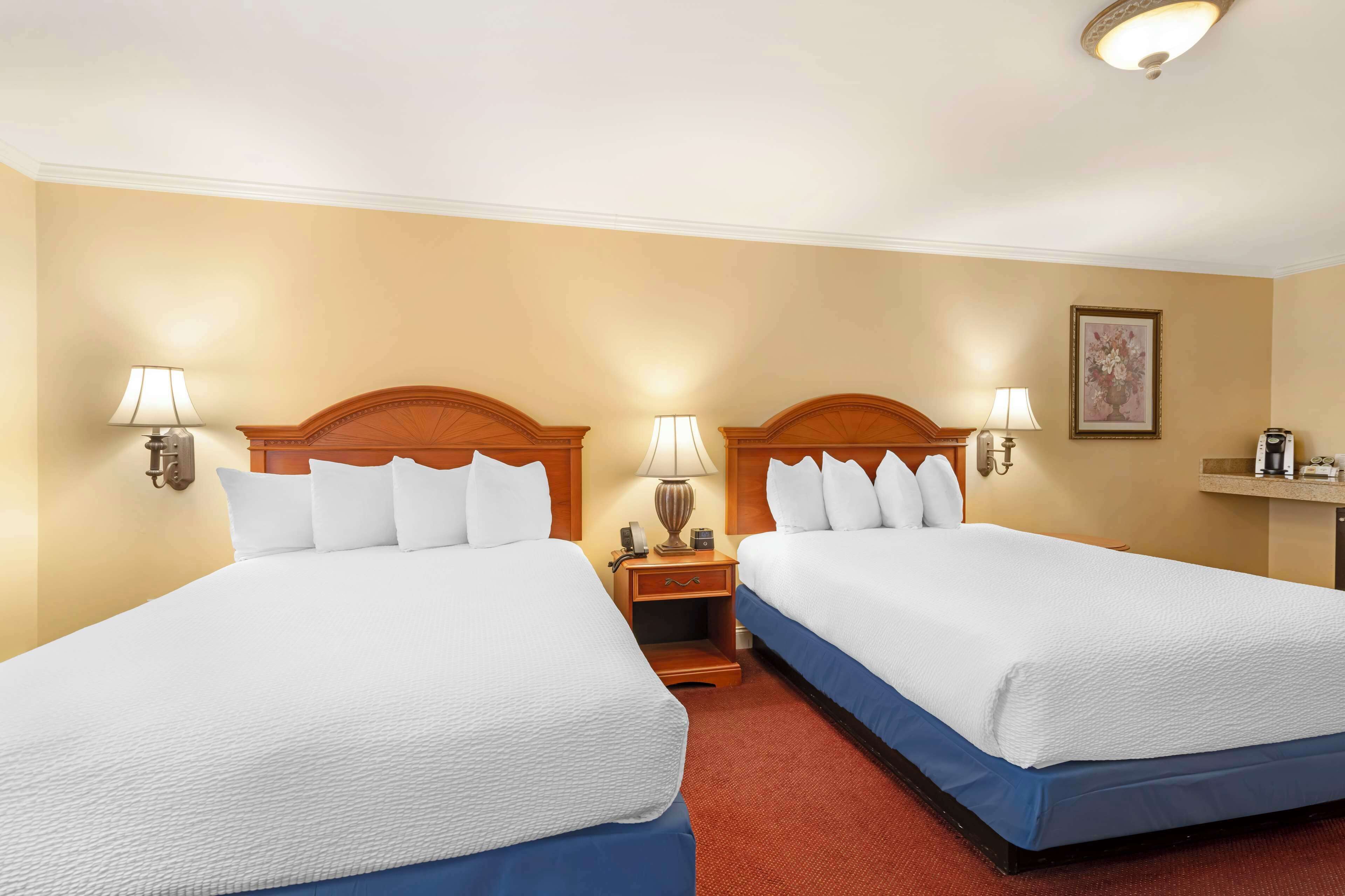 A hotel room with two beds, each with white bedding and a wooden headboard. There are bedside lamps, a nightstand with a lamp, and a framed picture on the wall.