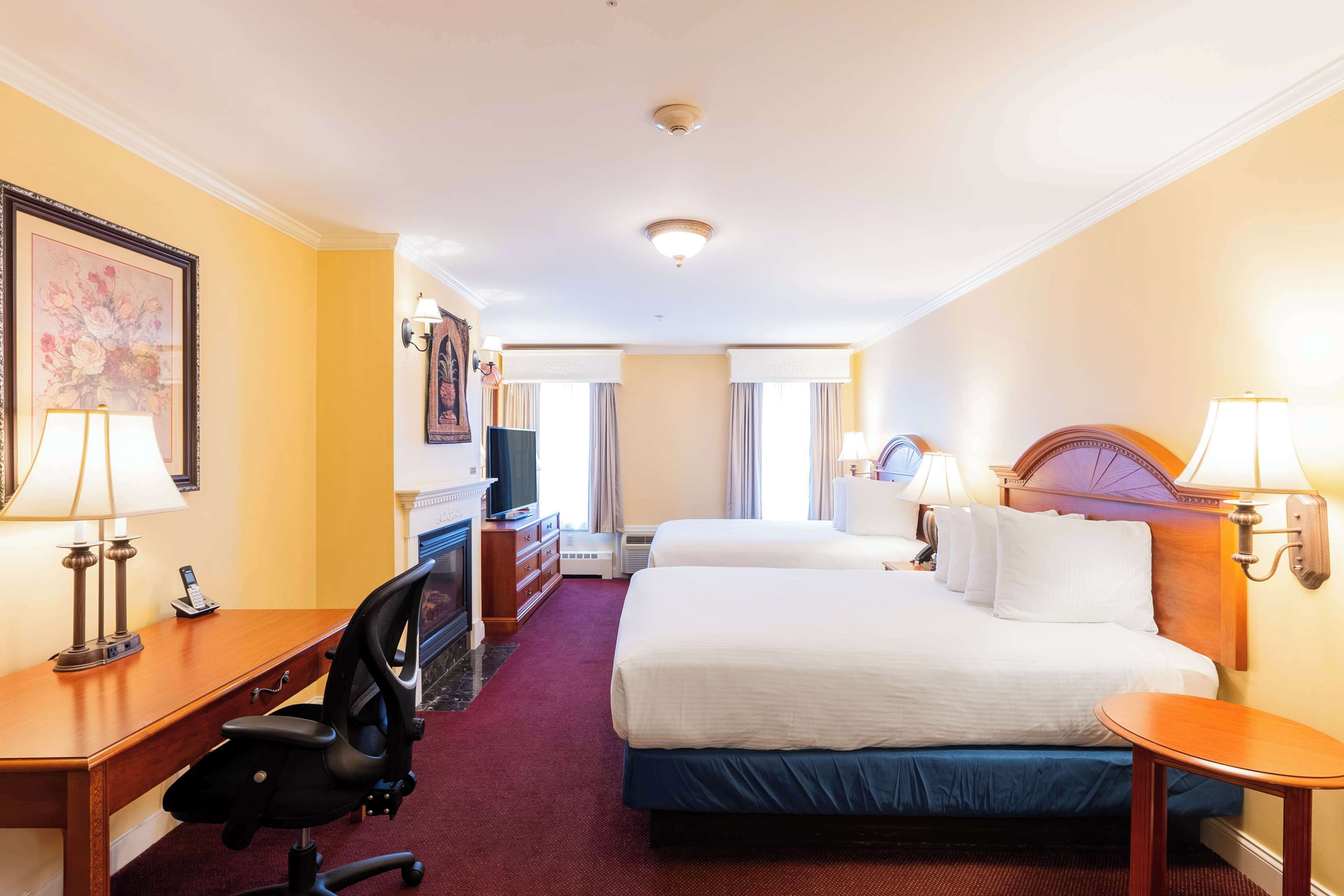 A hotel room with two beds, a desk with a chair, a fireplace, and a large window.