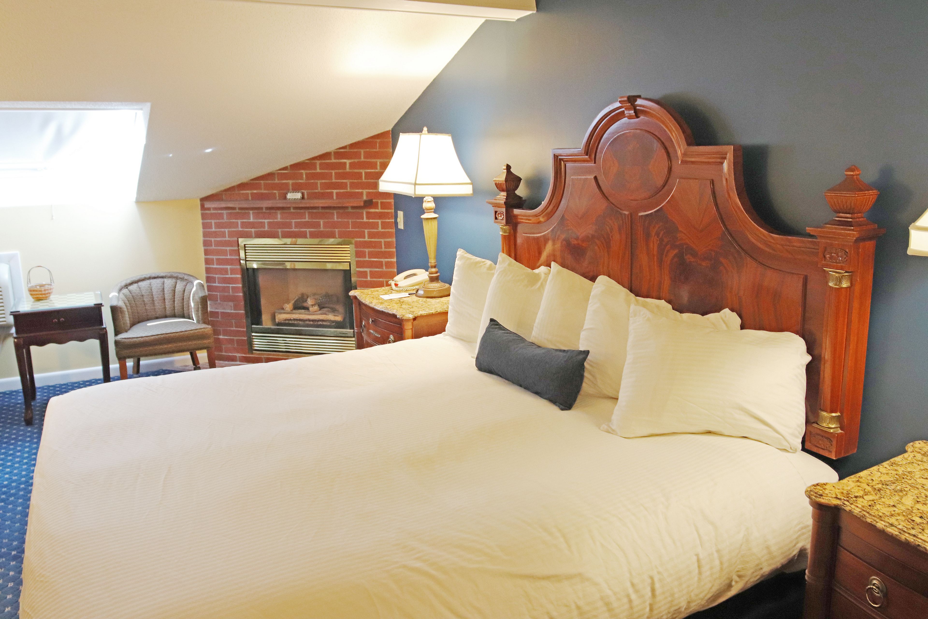 A hotel room with a large bed, a wooden headboard, a fireplace, a desk with a lamp, and a chair.