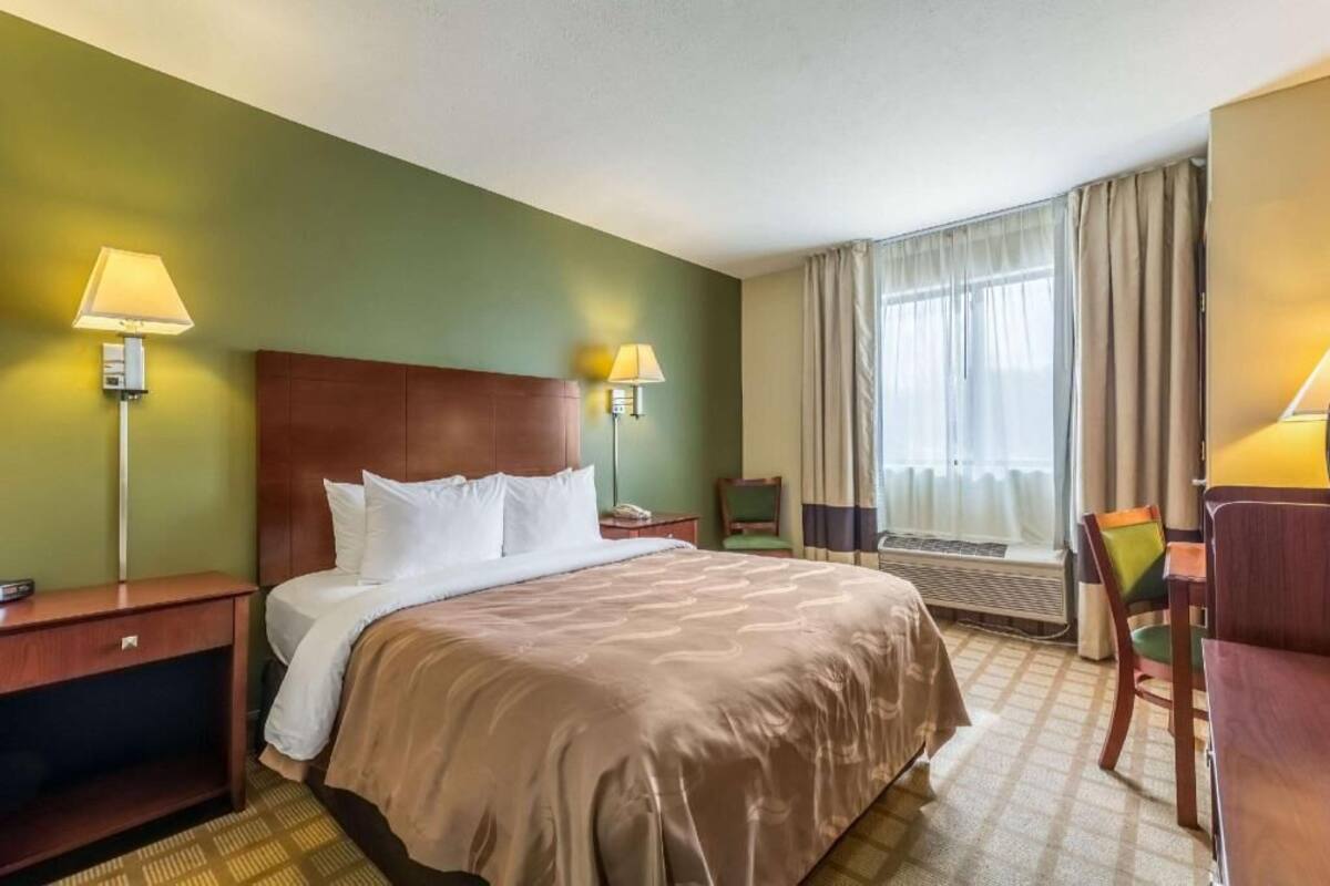 A hotel room with a bed, bedside tables, a desk, a chair, a window with curtains, and a lamp.