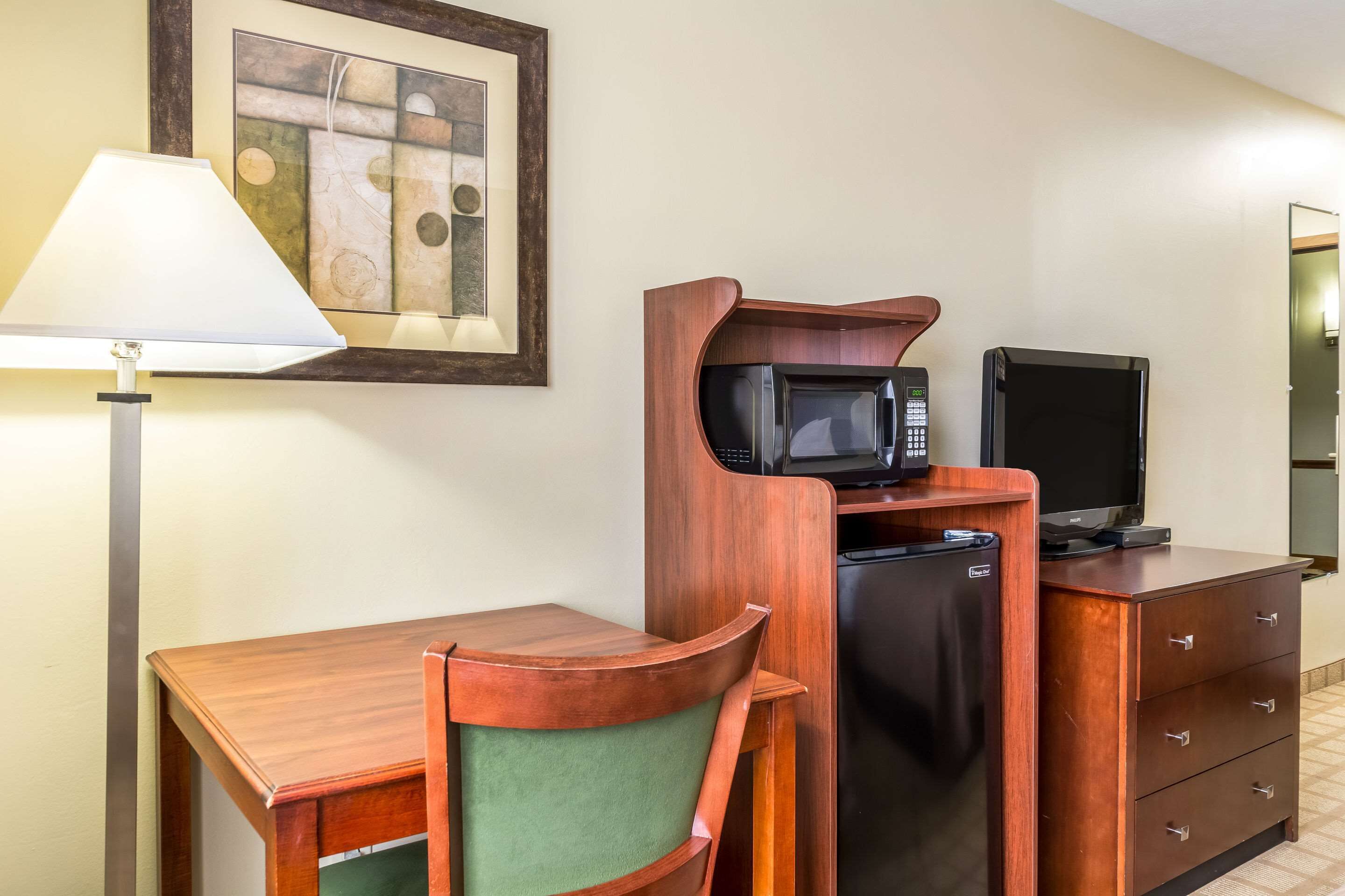 A compact room with a wooden desk, a green chair, a microwave, a television, a refrigerator, and a lamp.