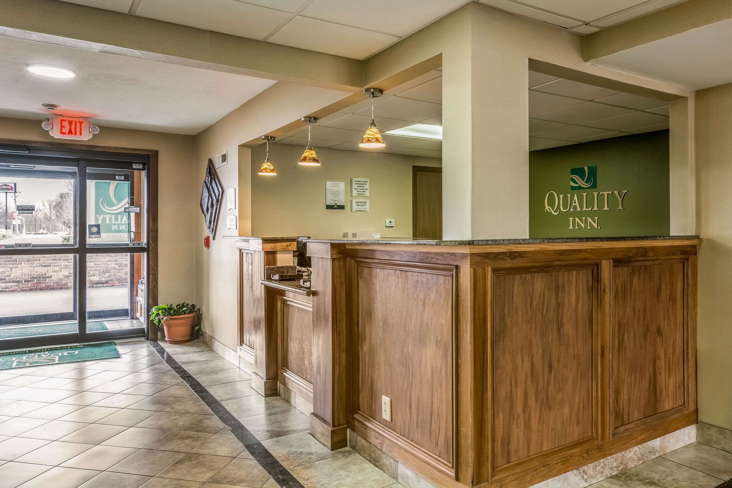 Zona de recepción de hotel con un mostrador de madera, una pared verde con el nombre del hotel y un piso de baldosas.