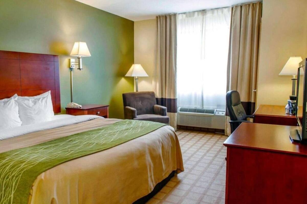 A hotel room with a bed, two chairs, a desk, a lamp, and an air conditioning unit.