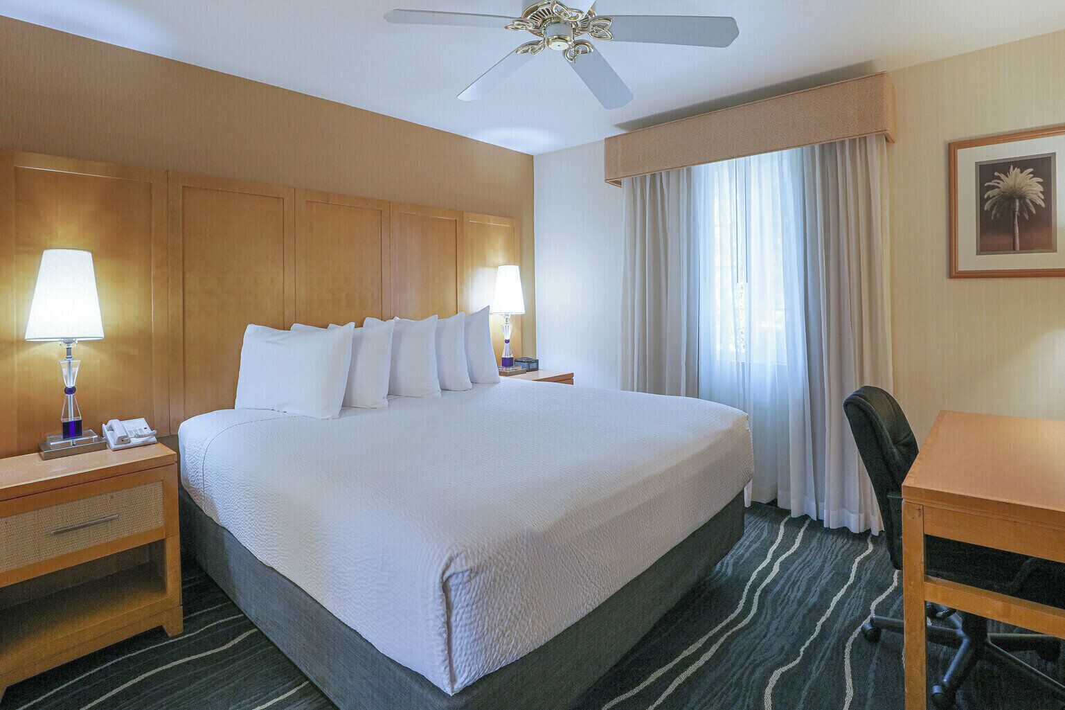 Habitación de hotel con una cama grande, dos mesitas de noche con lámparas, un escritorio, una silla, un ventilador de techo y una ventana con cortinas.