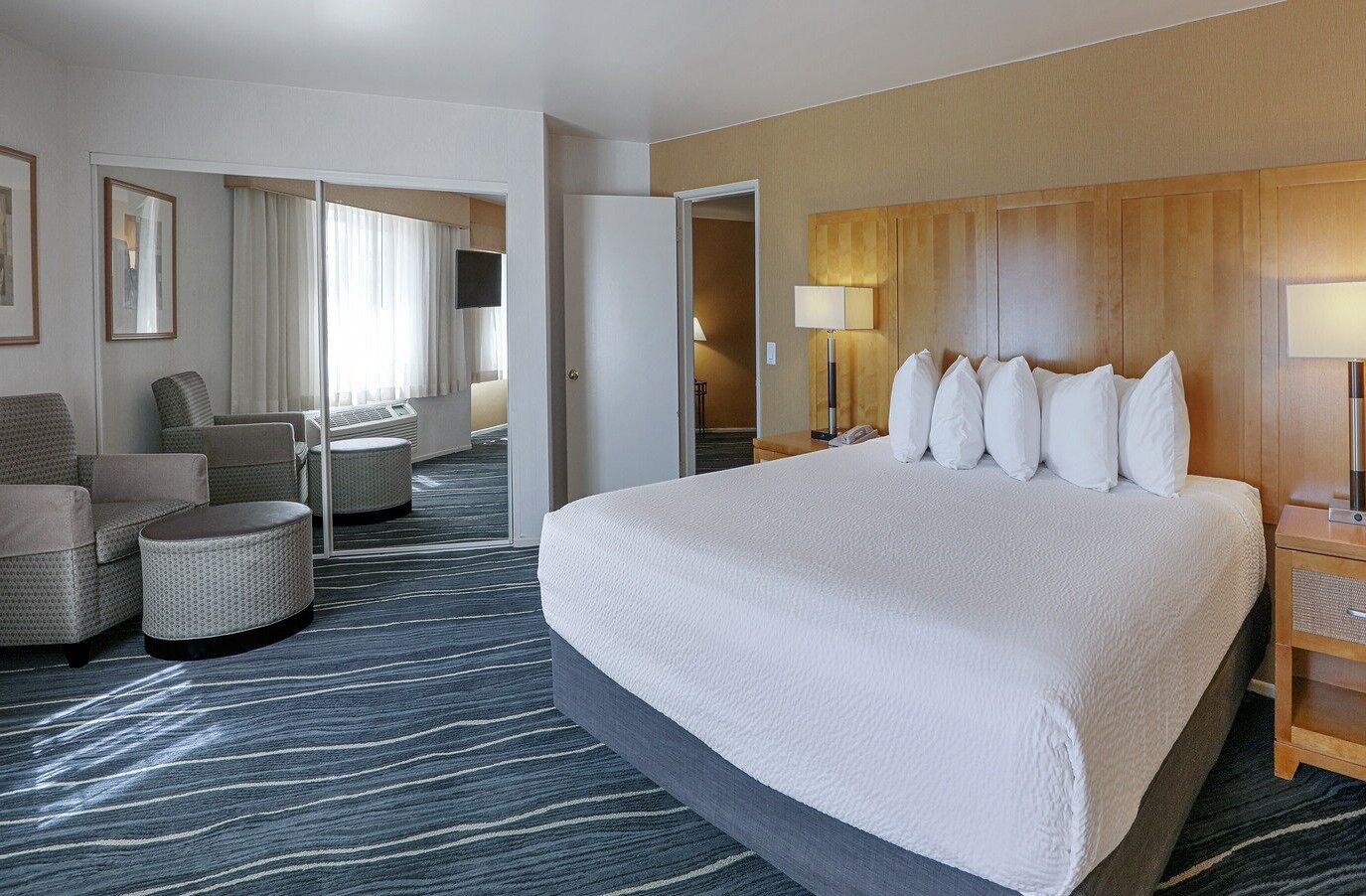 A hotel room with a large bed, a seating area with chairs and ottoman, a wooden headboard, and a view through a door.