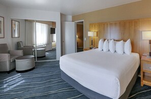 A hotel room with a large bed, a seating area with chairs and ottoman, a wooden headboard, and a view through a door. - Ramada by Wyndham Costa Mesa/Newport Beach (Costa Mesa)