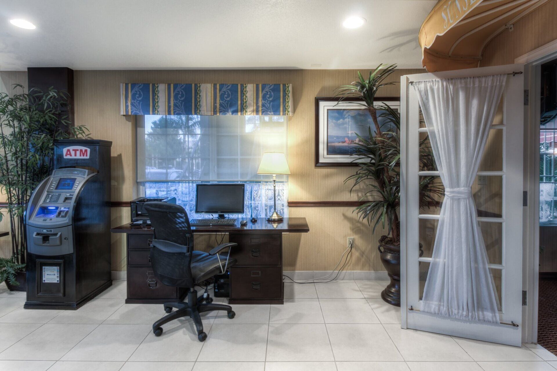 Una habitación de hotel con escritorio, cajero automático, computadora, silla y ventana con cortinas.