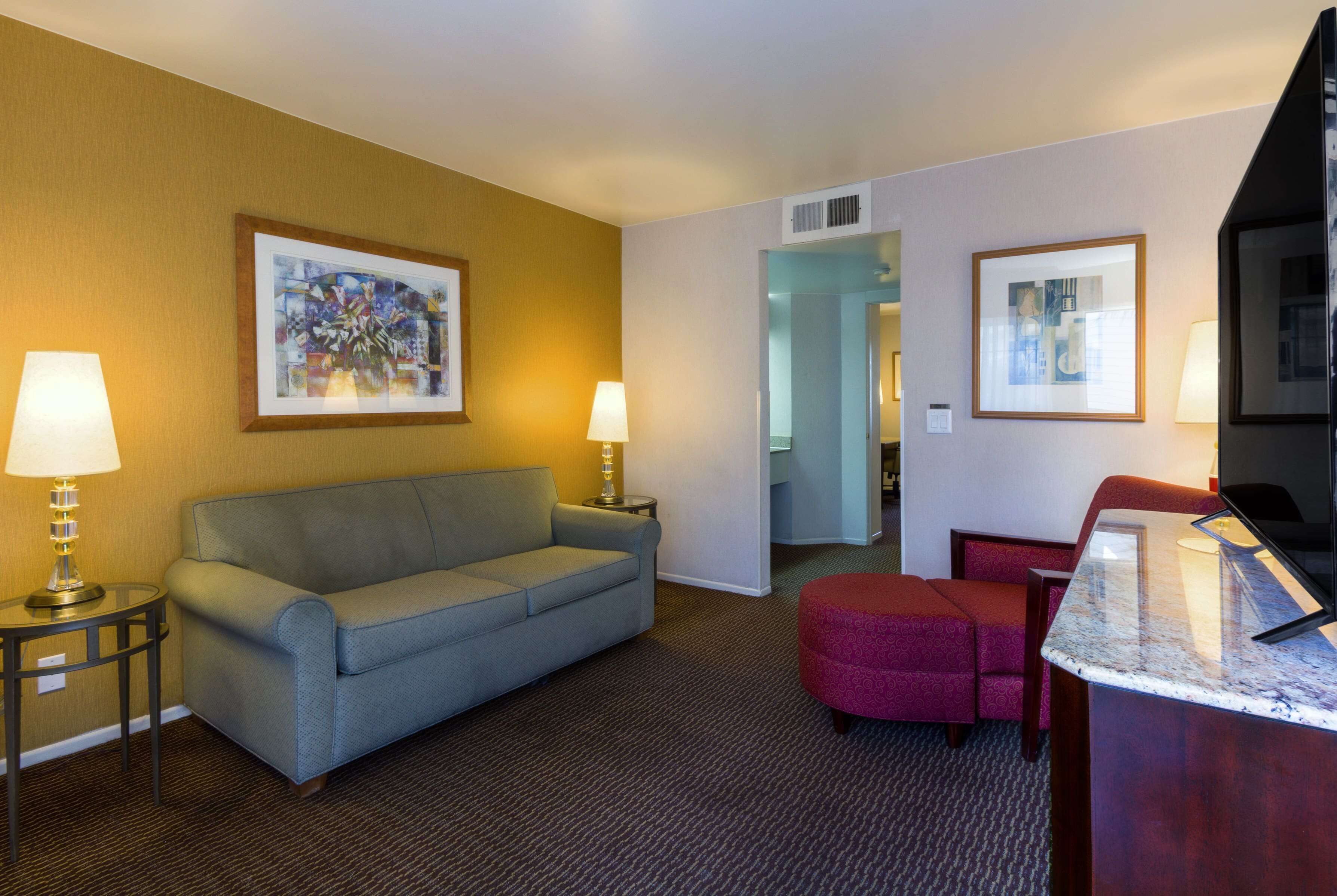 A hotel room with a sofa, a small table with a lamp, a red chair, and a desk with a computer.