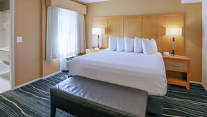 A hotel room with a large bed, two bedside tables with lamps, a bench, and a view of the hallway. - Ramada by Wyndham Costa Mesa/Newport Beach (Costa Mesa)
