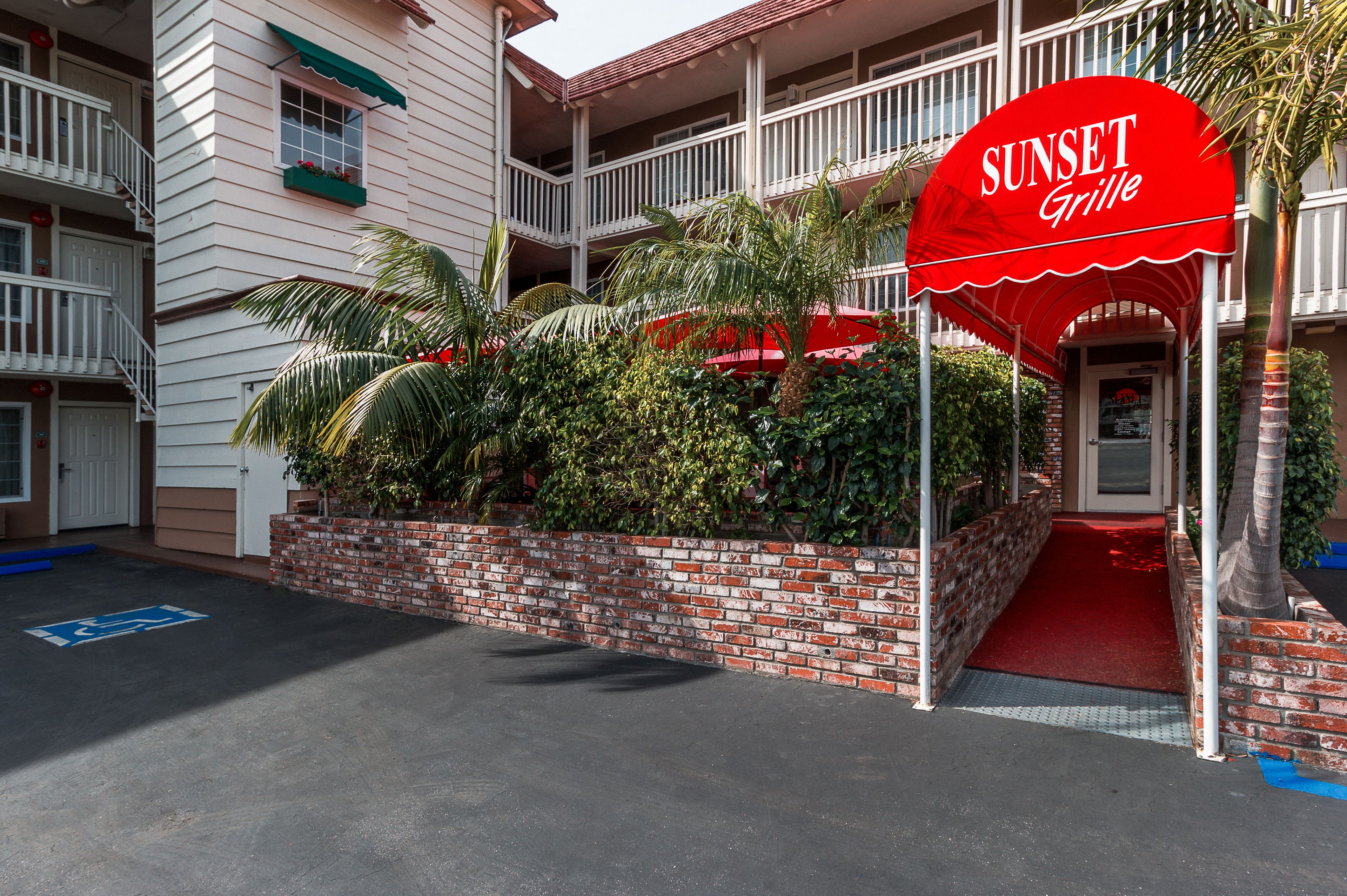 A hotel entrance with a red awning labeled "SUNSET Grille" and a brick wall with greenery.