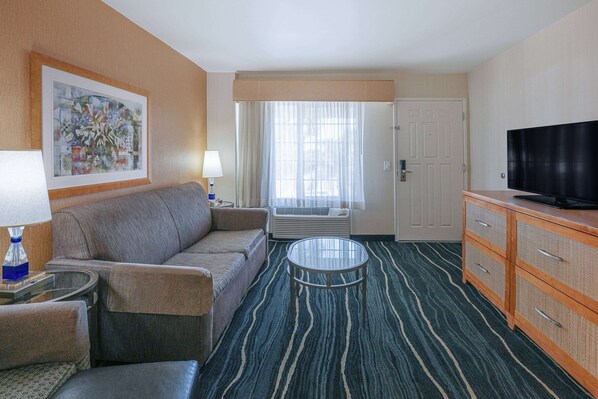 Habitación de hotel con sofá, una mesita, televisor, armario y un gran ventanal con cortinas.
