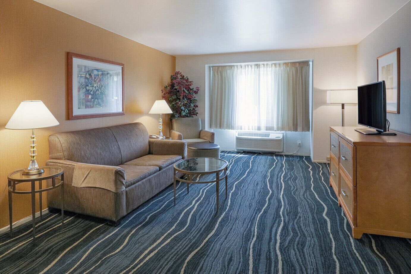 Habitación de hotel con sofá, dos mesitas de noche con lámparas, una pequeña mesa de centro, televisión sobre un mueble de madera y un amplio ventanal con cortinas.