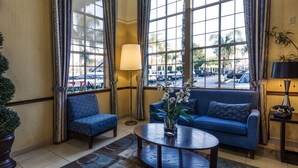 A hotel lobby with a blue sofa, a round coffee table with a vase of flowers, a floor lamp, and large windows with curtains. - Ramada by Wyndham Costa Mesa/Newport Beach (Costa Mesa)