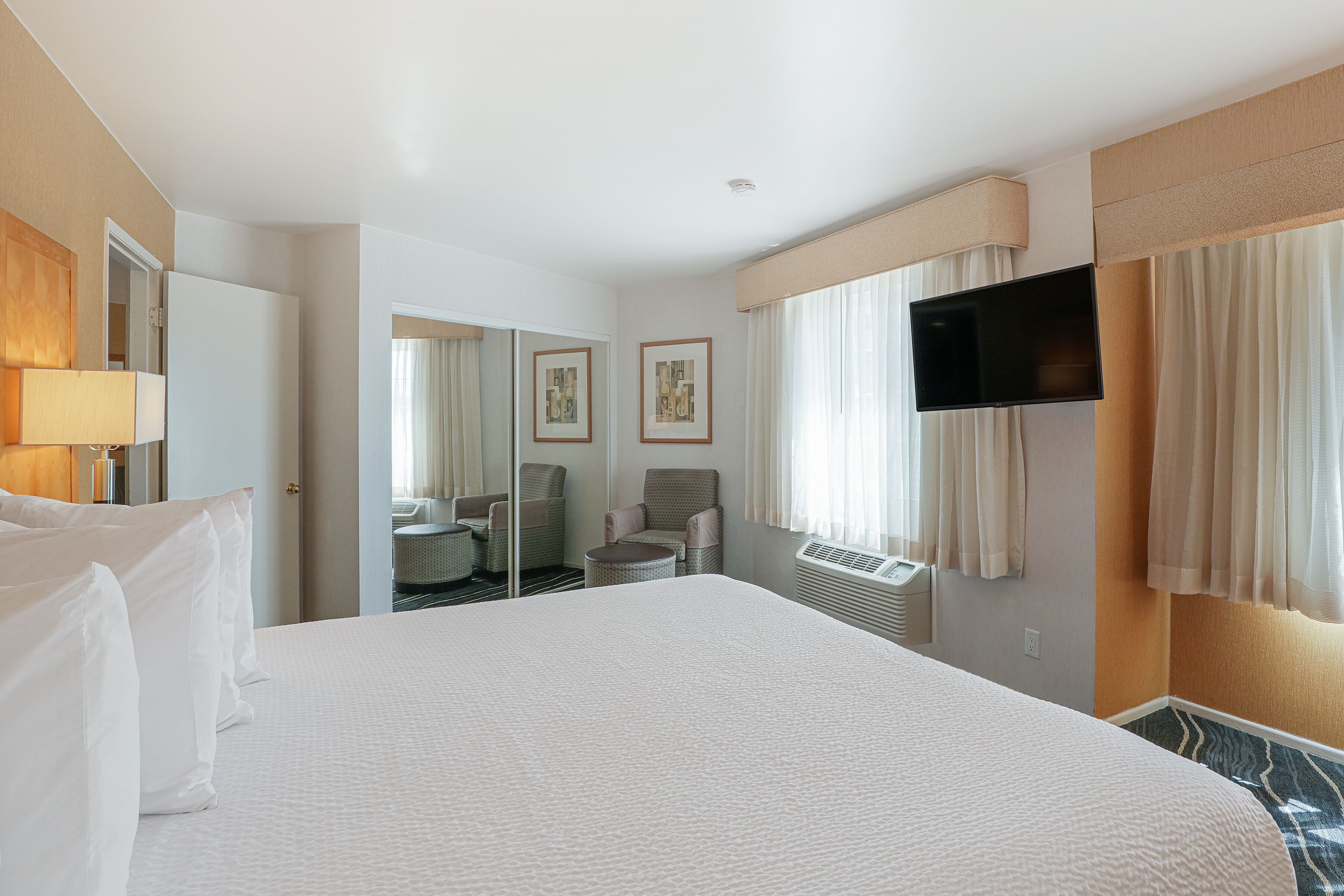 A hotel room with a large bed, a flat-screen TV mounted on the wall, a seating area with chairs, and a mirror.
