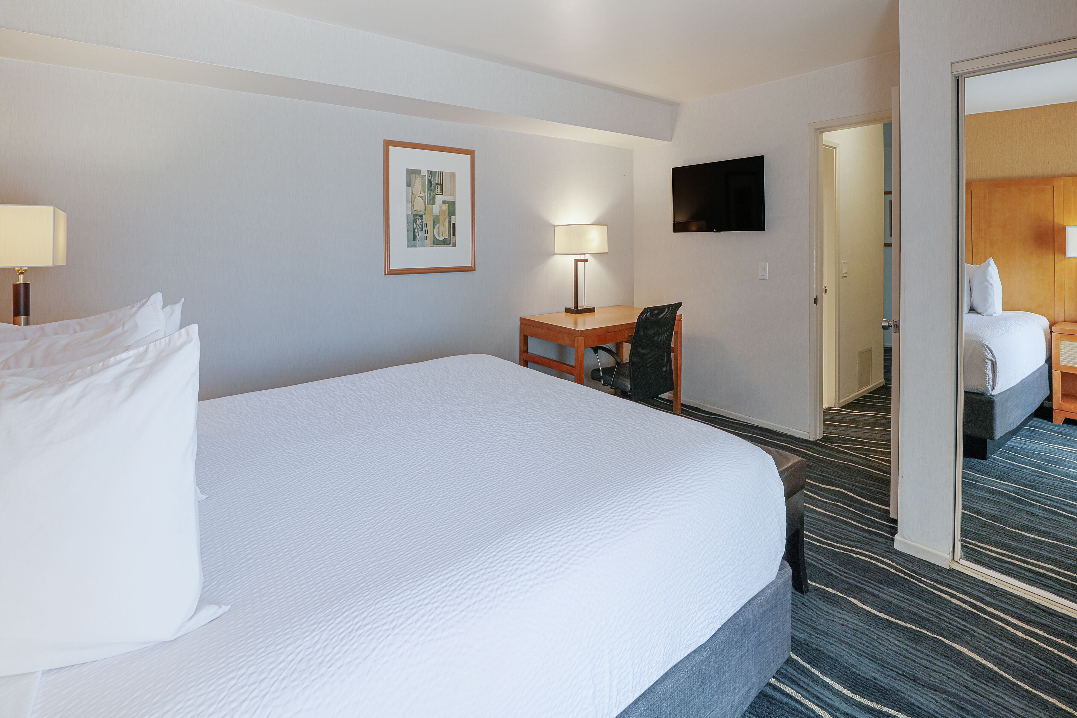 A hotel room with a large bed, a desk with a chair, a flat-screen TV mounted on the wall, and a framed picture above the desk.