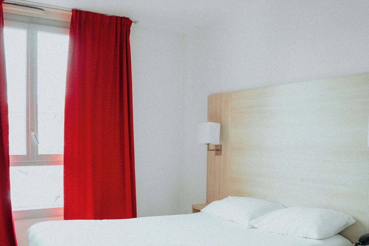 A hotel room with a bed, a bedside table, a lamp, and a window with red curtains.
