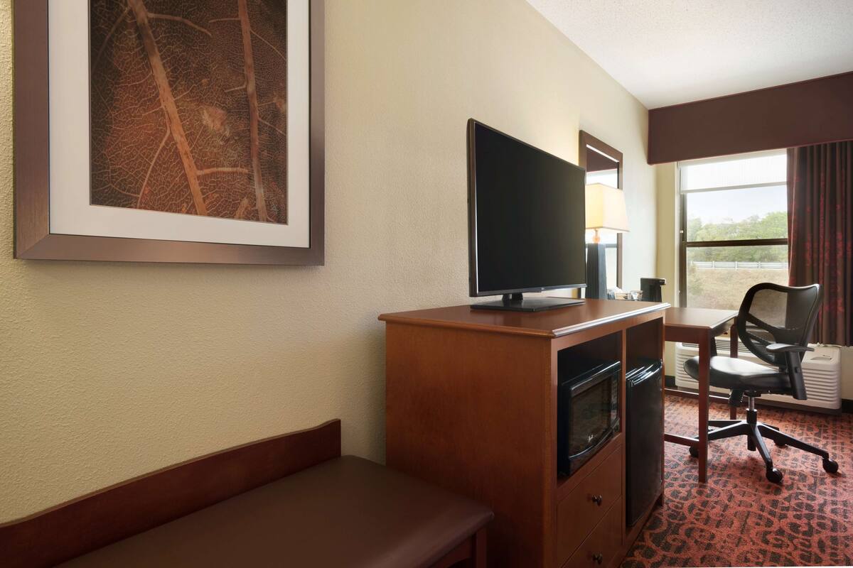 A hotel room with a desk, a television, a chair, a bench, and a large framed artwork on the wall.