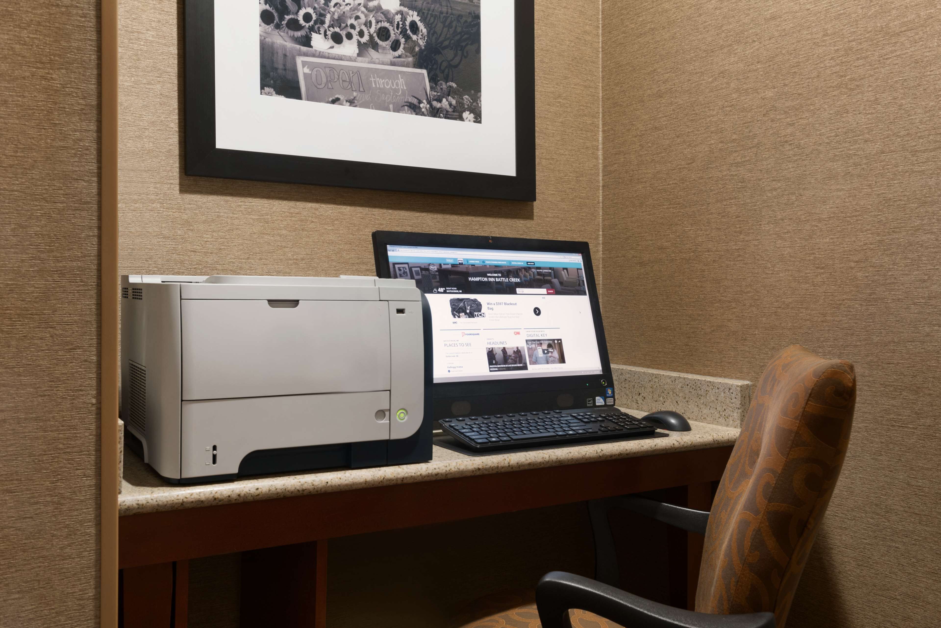 Un escritorio con una impresora y una computadora portátil, un cuadro en la pared y una silla.